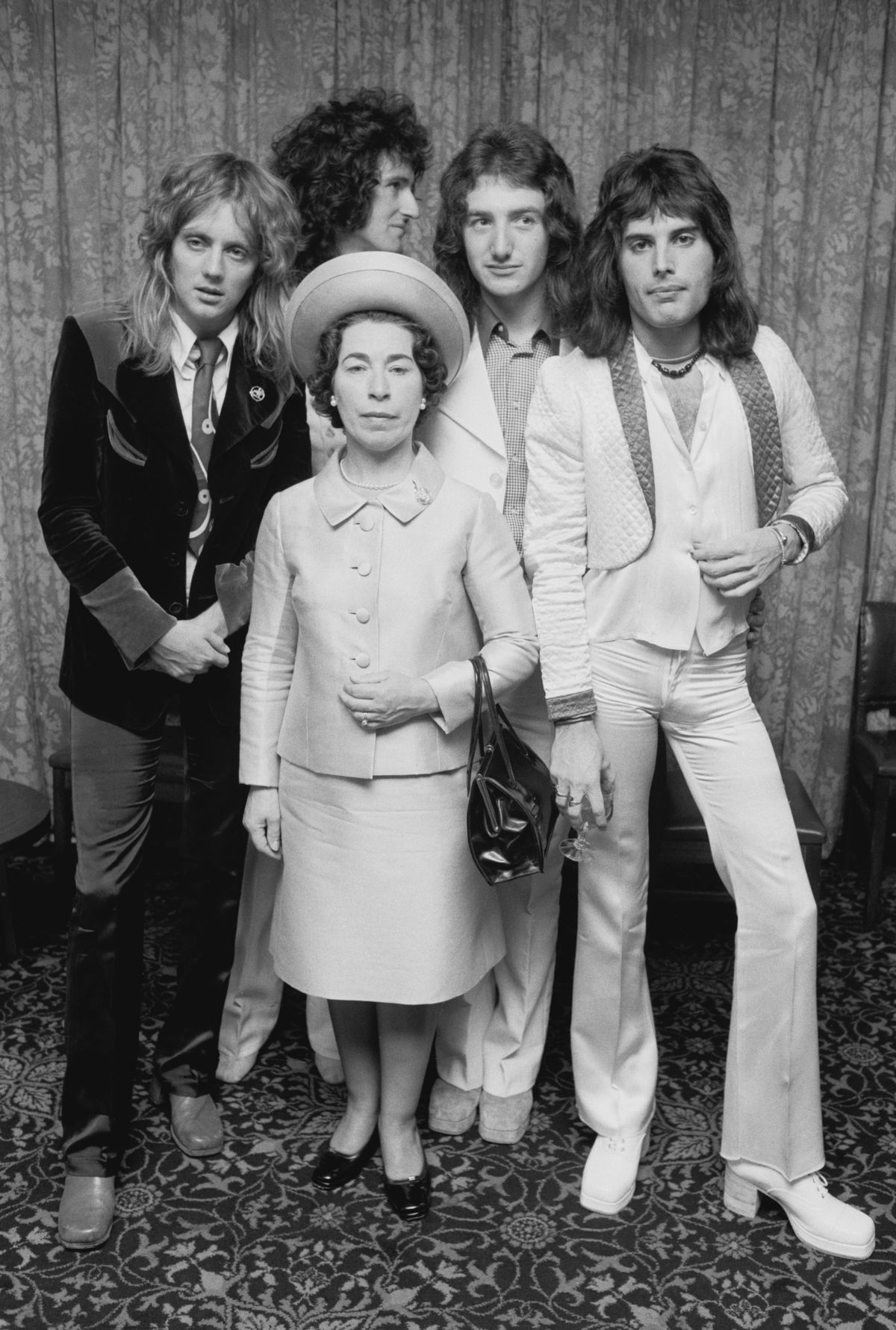 British rock band Queen posing with actress and Queen Elizabeth II look-alike, Jeannette Charles, September 1974. The group are (left to right) drummer Roger Taylor, guitarist Brian May, bassist John Deacon and singer Freddie Mercury (1946 - 1991). (Photo by Michael Putland/Getty Images)