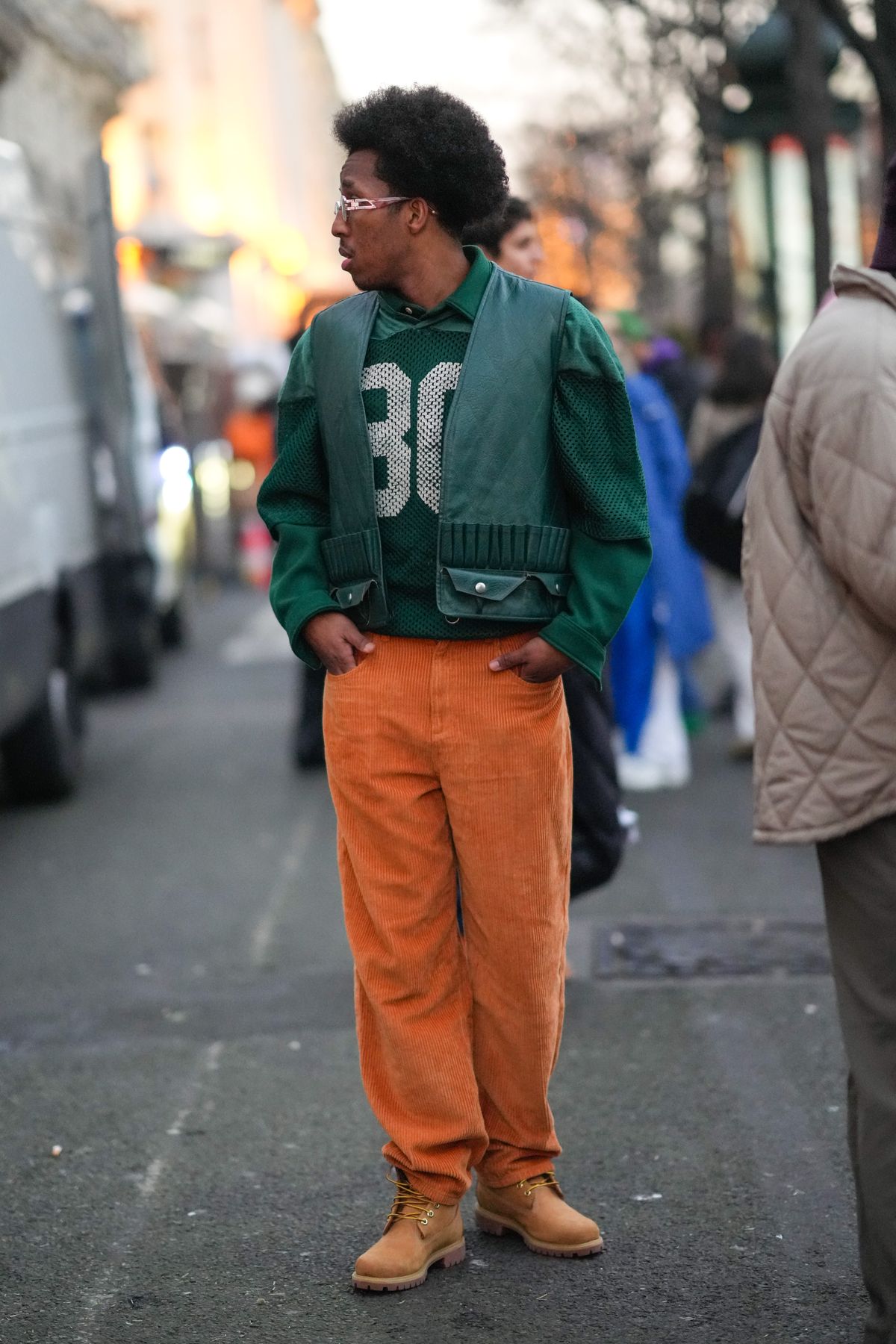 PARIS, FRANCE - JANUARY 21: A guest wears sunglasses, a dark green polo shirt, a dark green mesh sport t-shirt with a white 80 embroidered, a dark green shiny leather sleeveless jacket, orange ribbed velvet large pants, brown ankle boots from Timberland, outside Jil Sander, during Paris Fashion Week - Menswear F/W 2022-2023, on January 21, 2022 in Paris, France. (Photo by Edward Berthelot/Getty Images)