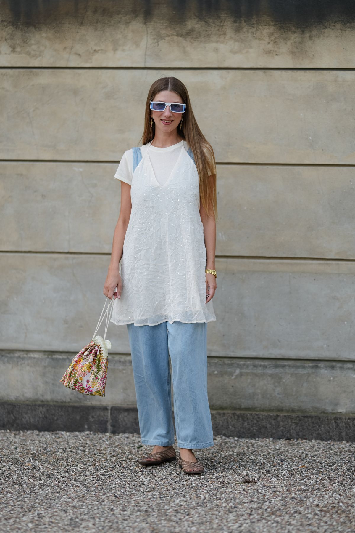 COPENHAGEN, DENMARK - AUGUST 06: A guest wears rectangular blue sunglasses and long, straight hair. A white short-sleeved t-shirt is layered under a white sleeveless dress featuring textured embroidery. Light blue wide-leg jeans are worn below. Accessories include a small floral-patterned drawstring bag and a gold wristwatch. Brown woven flat shoes complete the outfit, outside Munthe, during Copenhagen Fashion Week, on August 06, 2025 in Copenhagen, Denmark (Photo by Edward Berthelot/Getty Images)