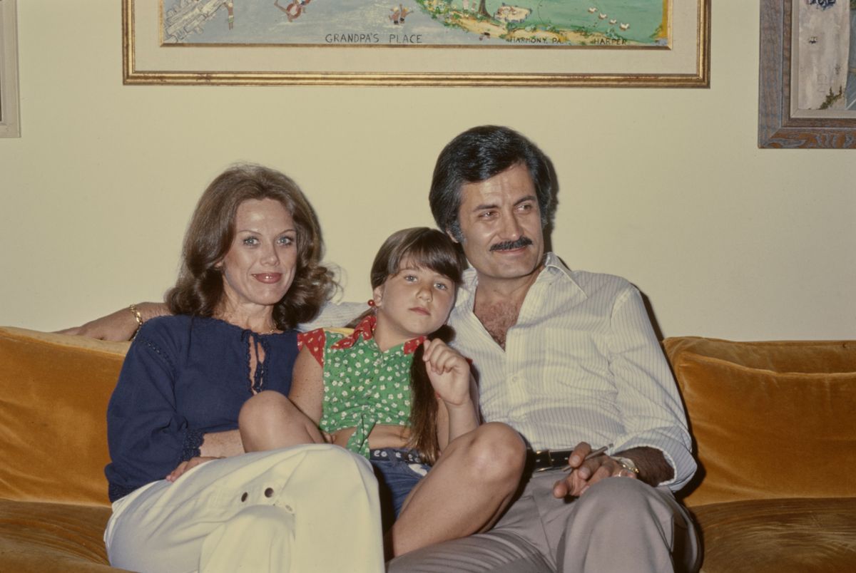 American actress Nancy Dow, wearing a dark blue blouse, sitting on a sofa with her daughter, American actress Jennifer Aniston, wearing a red and green polka dot tie-front shirt, and her husband Greek-born American actor John Aniston, in a striped white shirt, open at the collar, and holds a lit cigarette, at the family home in the Sherman Oaks neighbourhood of Los Angeles, California, 1975. (Photo by Michael Ochs Archives/Getty Images)