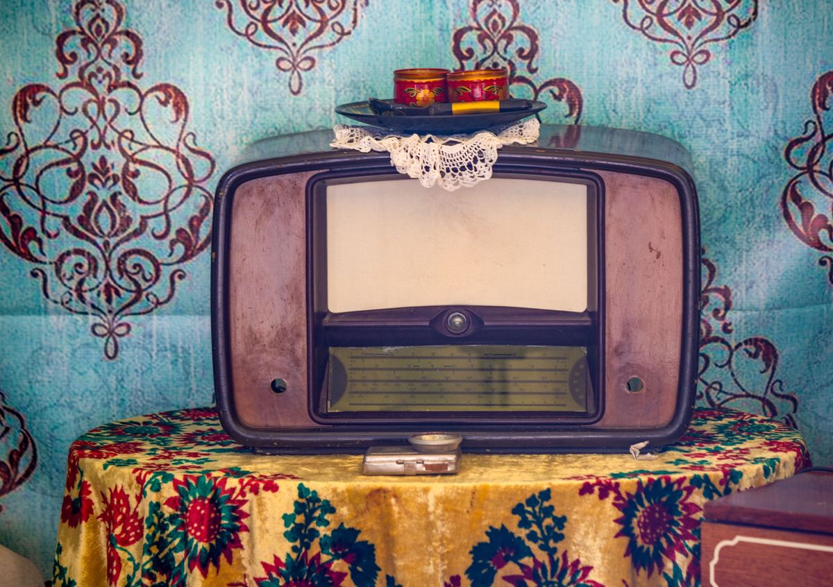 A vintage radio sits on a table next to a floral tablecloth. The radio is old and has a lace doily on top of it. The tablecloth is colorful and has a floral pattern. The scene gives off a nostalgic