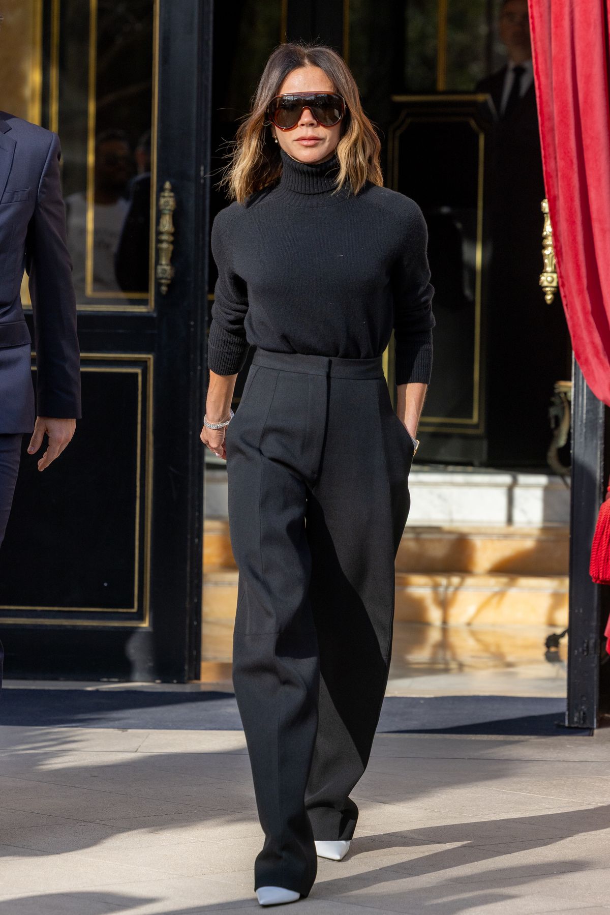 Victoria Beckham egy teljesen fekete, garbós szettben, napszemüveggel és egy fehér magassarkúval
