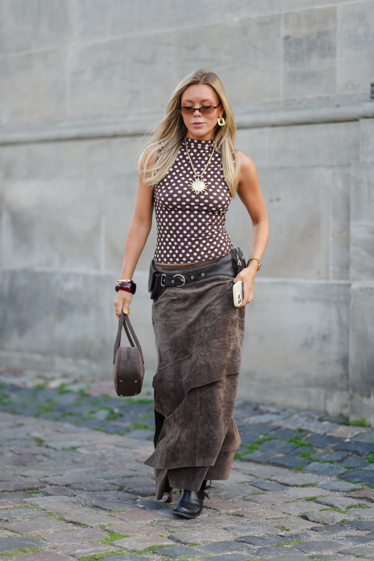 COPENHAGEN, DENMARK - AUGUST 06: A guest wears sunglasses with tinted lenses and a sleeveless, high-neck top featuring a brown and white polka dot pattern. Hair is styled straight and loose. Large hoop earrings and a gold sun pendant necklace are visible. A dark brown, layered suede skirt is paired with a wide black belt. Accessories include a brown structured handbag and a smartphone held in hand. Black ankle boots complete the ensemble, outside The Garment, during Copenhagen Fashion Week, on August 06, 2025 in Copenhagen, Denmark (Photo by Edward Berthelot/Getty Images)