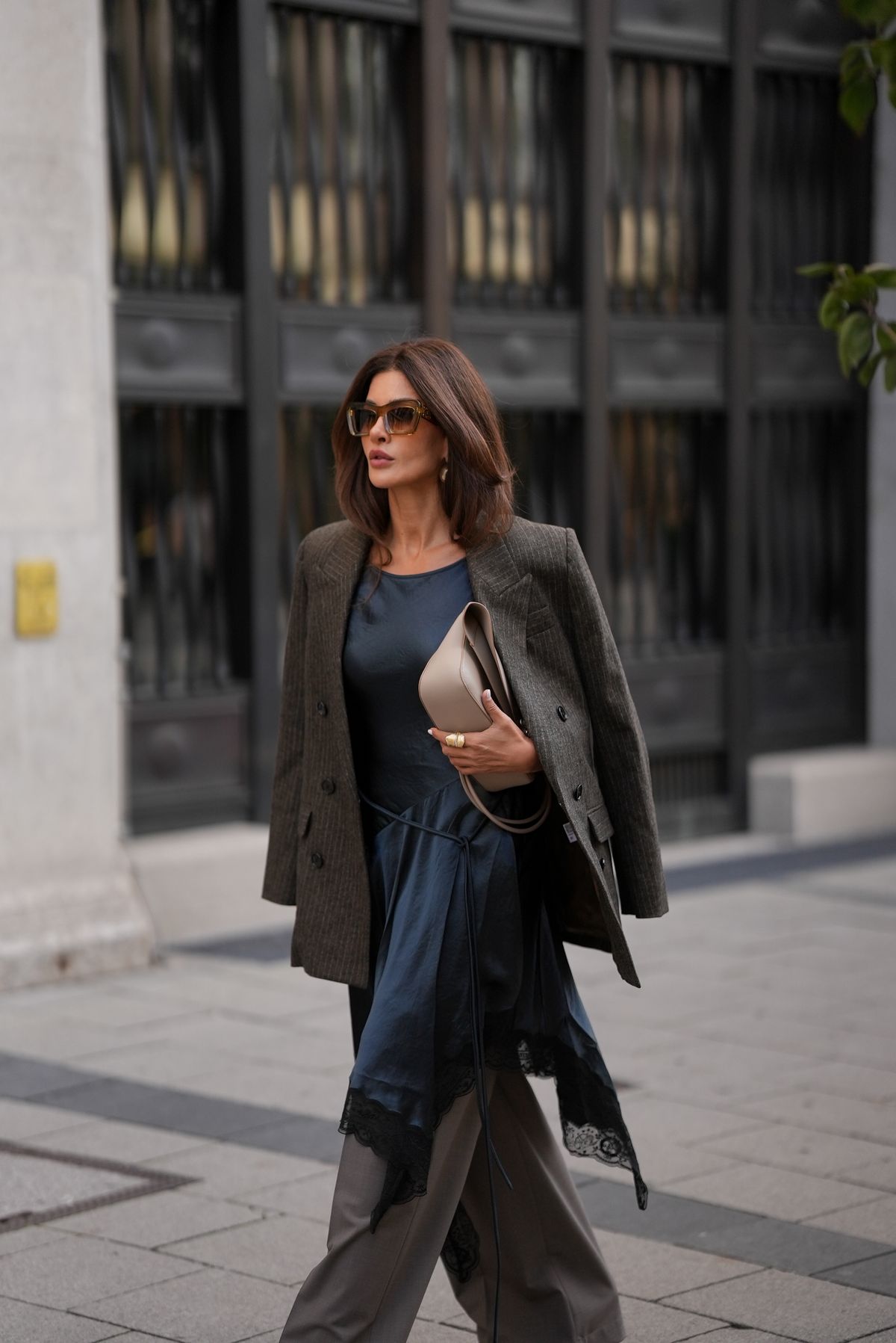 MUNICH, GERMANY - SEPTEMBER 3: Füsun Lindner is seen wearing a dark grey, double-breasted blazer draped casually over the shoulders from Isabel Marant Etoile; a flowing navy silk dress with an asymmetrical handkerchief hem, finished with black lace trim from Acne; grey tailored suit trousers in a straight cut from Acne; a light beige leather handbag with gold hardware and minimal branding from Loewe; oversized brown-tinted sunglasses with chunky frames from Loewe; her brown hair is worn in a straight bob with a center part on September 3, 2025 in Munich, Germany. (Photo by Moritz Scholz/Getty Images)