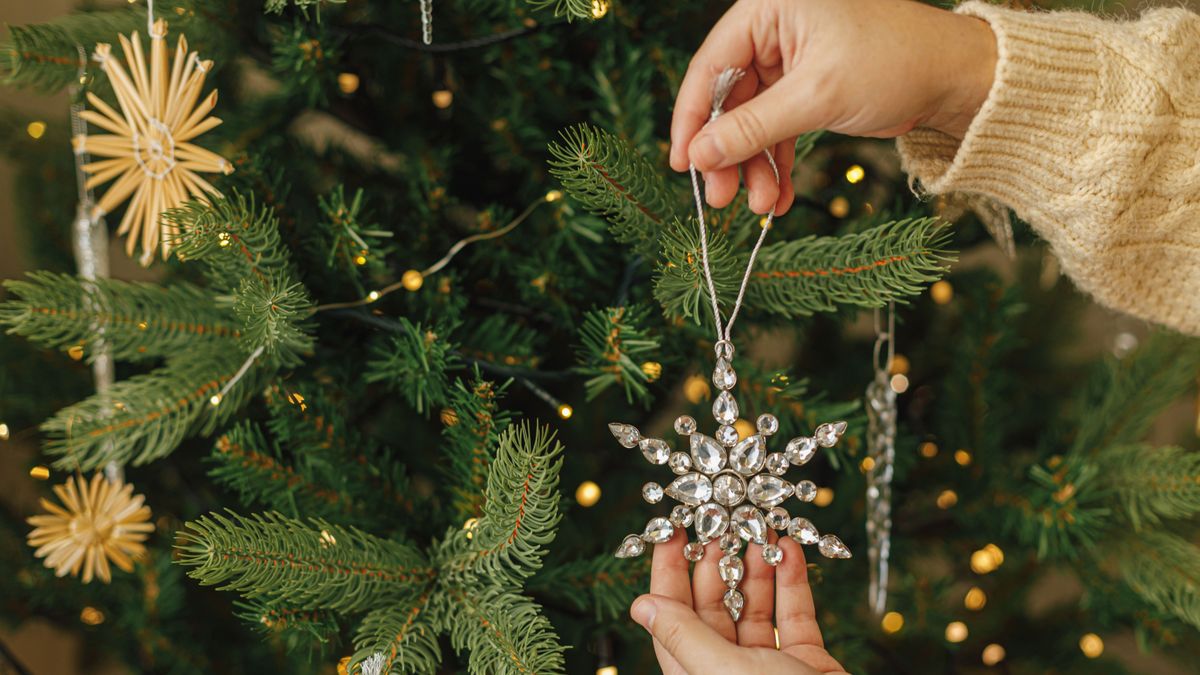 Hands in knitted sweater hanging stylish Christmas ornament on christmas tree with golden lights. Decorating christmas tree with vintage glass snowflake