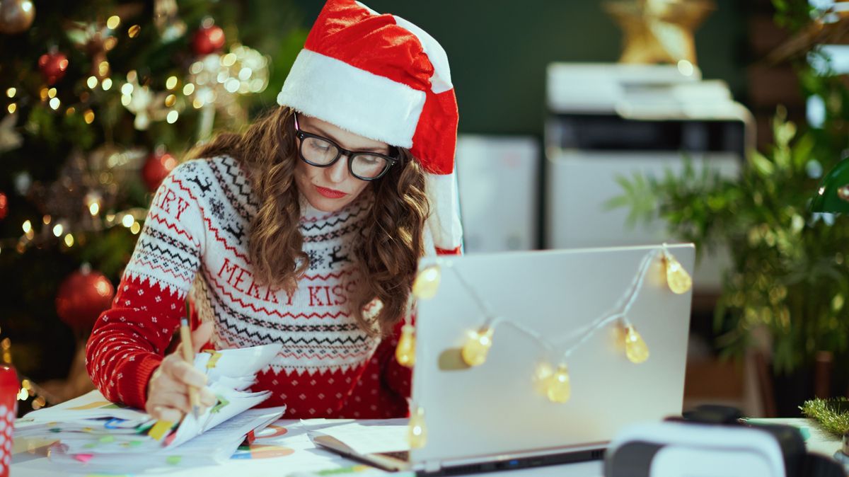 Christmas time. tired stylish middle aged small business owner woman in santa hat in eyeglasses in red Christmas sweater with documents and laptop working in modern green office with Christmas tree.