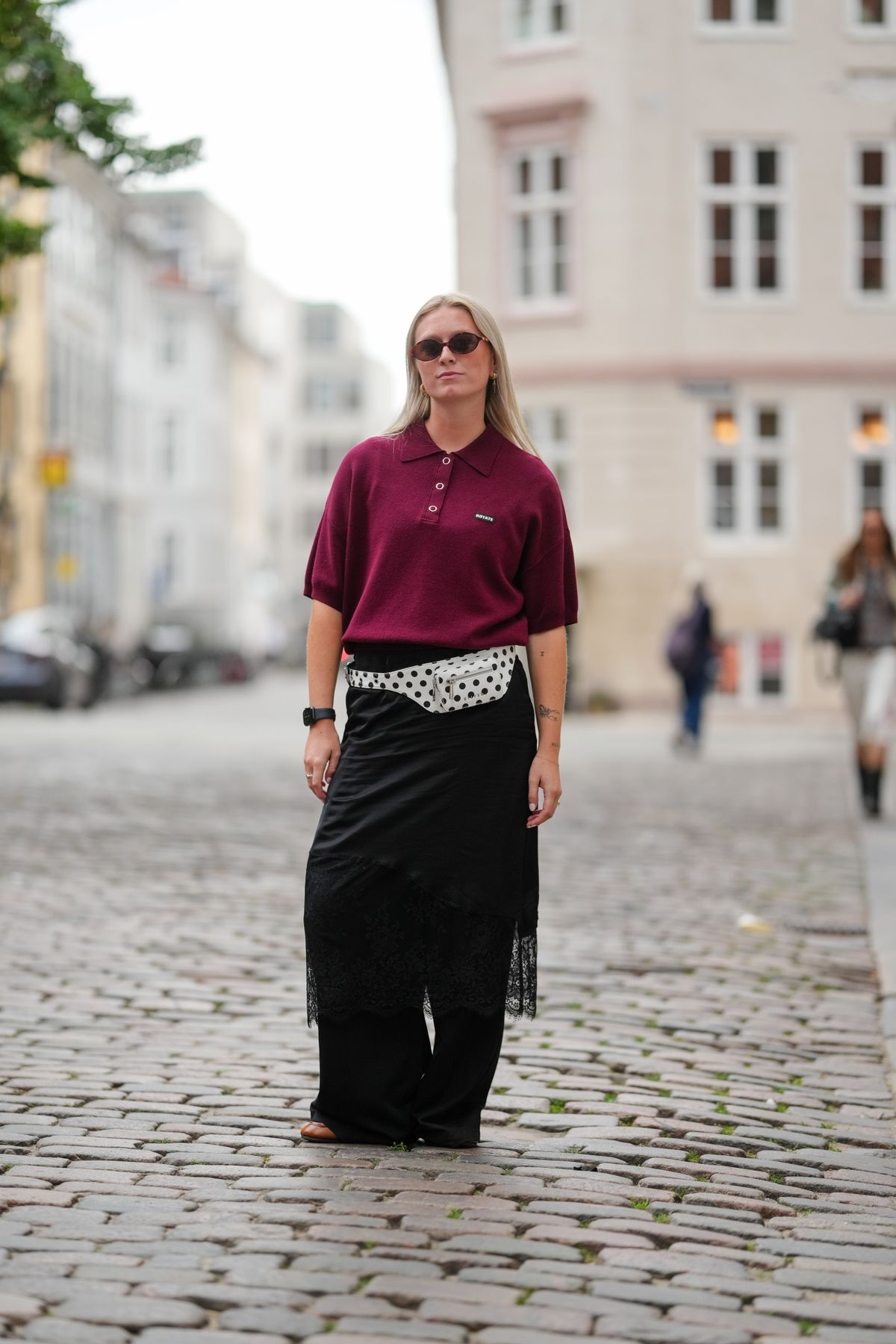 COPENHAGEN, DENMARK - AUGUST 07: A guest wears long straight blonde hair parted in the center, round brown sunglasses, and small gold hoop earrings. A burgundy knit polo shirt with a pointed collar, short sleeves, a four-button placket, and a small chest logo patch is styled with a white belt bag in black polka dots with a zip closure at the waist. A black wrap-style midi skirt with a sheer lace hem is layered over wide-leg black trousers. Brown leather flats are worn, with a black smartwatch on the left wrist and a silver-tone ring, posing, outside Nicklas Skovgaard, during Copenhagen Fashion Week, on August 07, 2025 in Copenhagen, Denmark (Photo by Edward Berthelot/Getty Images)