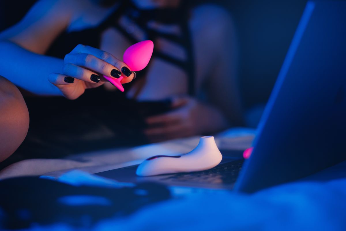 Beautiful woman lies in bed holding clitoral vibrator and watching porn on laptop, neon color. Women has Internet sex chat online.