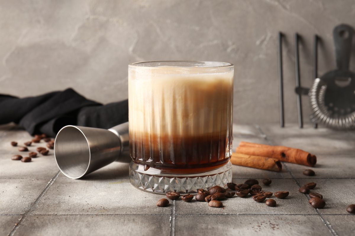Glass of White Russian cocktail with cinnamon and coffee beans on grey tile table