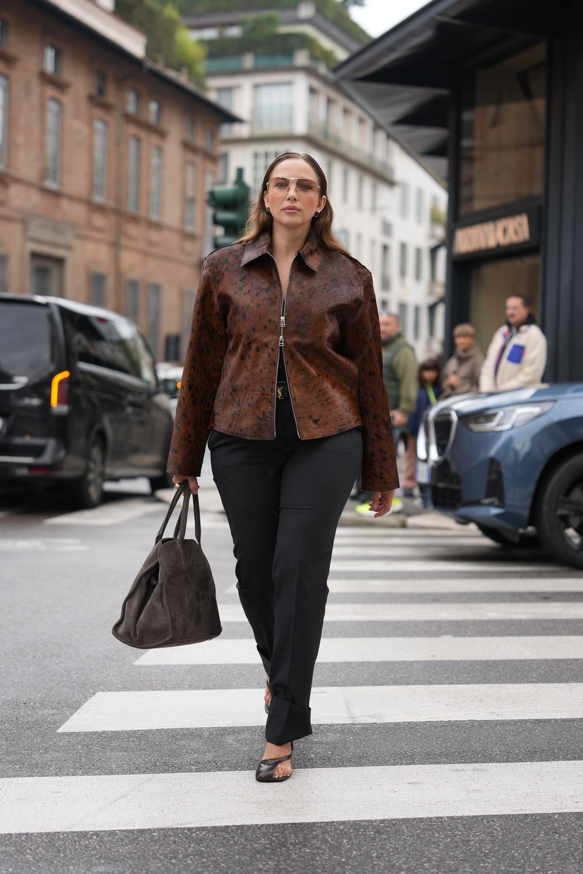 MILAN, ITALY - SEPTEMBER 27: A guest wears long brown hair worn straight with a center part, brown-tinted aviator sunglasses, small gold hoop earrings, grey suede tote bag, brown spotted leather zip-front jacket with a pointed collar, black tailored cuffed trousers, black textured open-toe mule shoes with a low heel, outside Ferragamo, during Milan Fashion Week - Womenswear Spring/Summer 2026, on September 27, 2025 in Milan, Italy (Photo by Edward Berthelot/Getty Images)