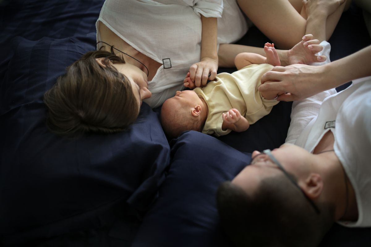 Newborn between parents on a dark sheet, dad and mom with the baby on the bed, top view. Parenthood and care concept, traditional Caucasian young family