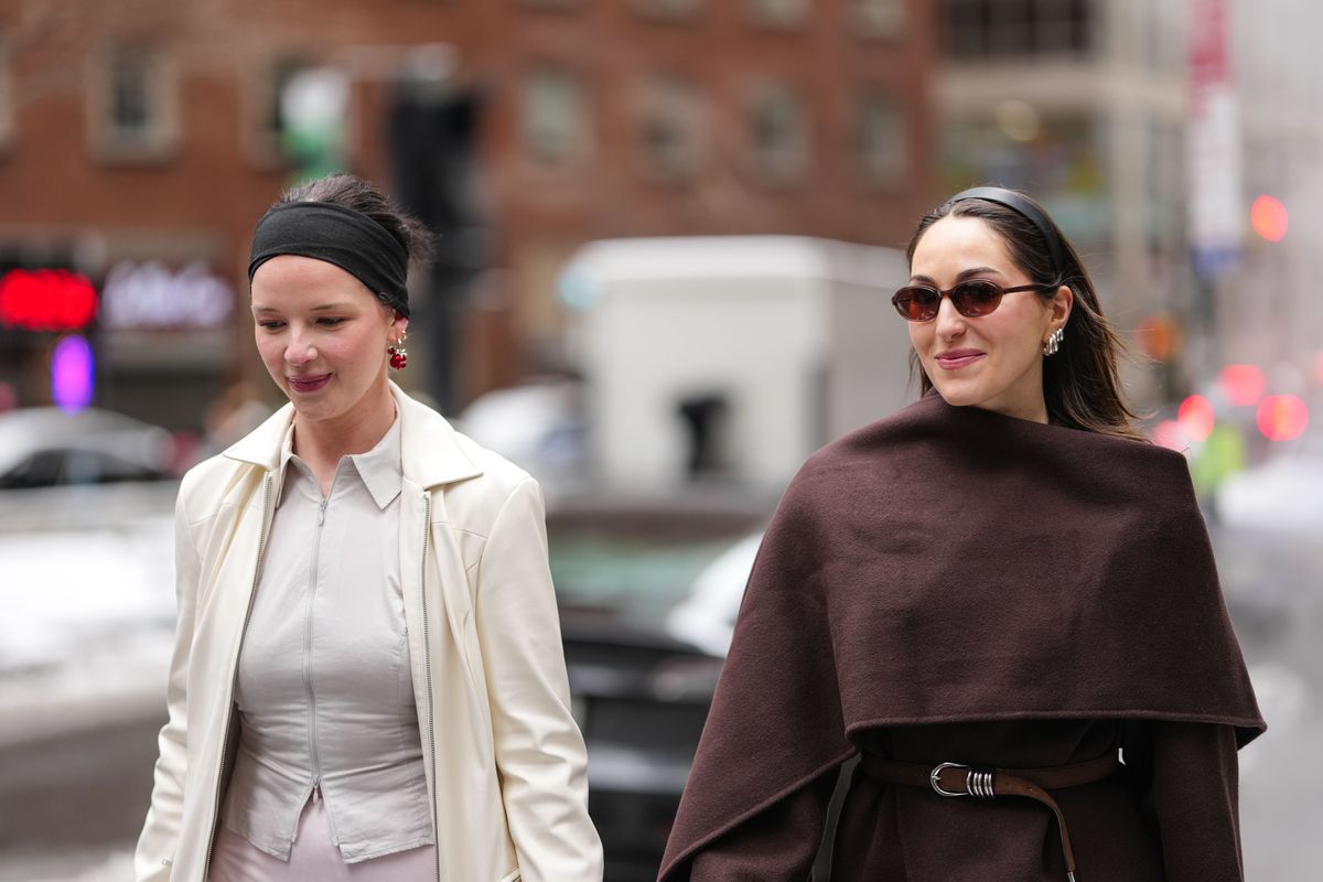 NEW YORK, NEW YORK - FEBRUARY 09: A guest wears dark brown sunglasses, silver earrings, dark brown large scarf, burgundy bordeaux wool coat, dark brown leather belt, shiny black leather bag, bag charms. A guest wears black headband, gold earrings, light gray zipped tailored jacket, cream coat, light pink sheer mesh midi skirt, outside Sandy Liang, during the New York Fashion week Fall/Winter 2025 on February 9, 2025 in New York, New York. (Photo by Edward Berthelot/Getty Images)