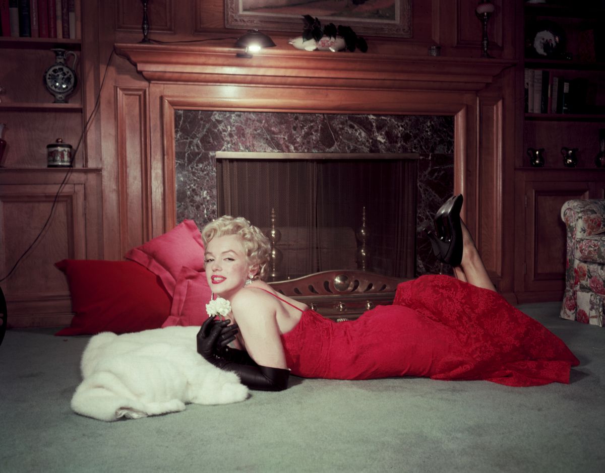 1955:  EXCLUSIVE Portrait of American actor Marilyn Monroe (1926 - 1962) as she holds a white carnation while lying on her stomach in front of a fireplace. Monroe wears a red evening dress, long black gloves, and black high heels.  (Photo by Gene Lester/Getty Images)