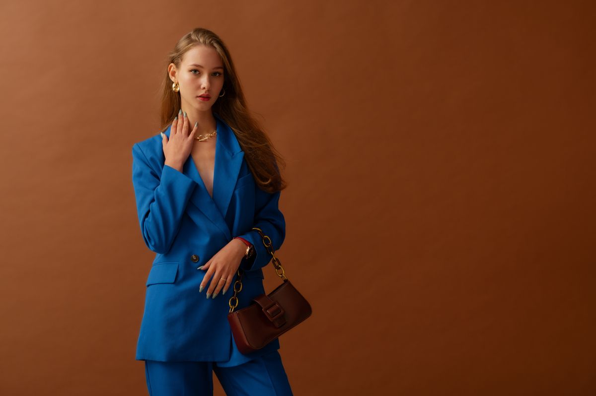 Fashionable confident woman wearing trendy blue suit blazer, trousers, holding small leather bag, posing in studio, on brown backdrop. Copy, empty space for text