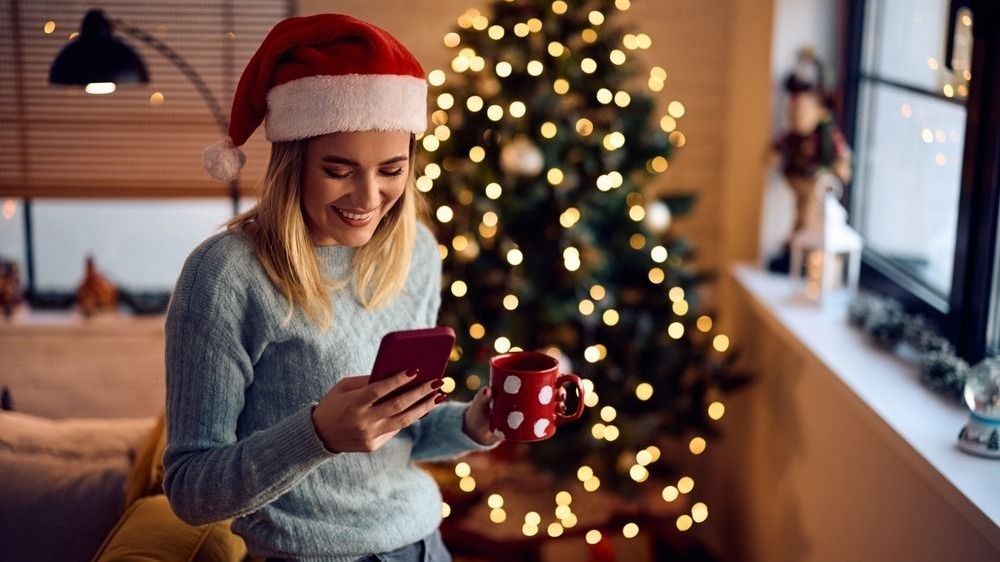 Fiatal nő kávétiszik, mögötte karácsonyfa, kezében telefon.