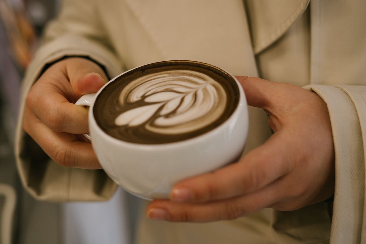 A close-up image showing a woman's hands holding a cup of hōjicha latte with latte art on top. The roasted green tea gives the drink a warm, earthy tone, contrasting beautifully with the creamy milk foam. Captured in soft, natural light, the image evokes a sense of calm and comfort, highlighting modern Japanese tea culture