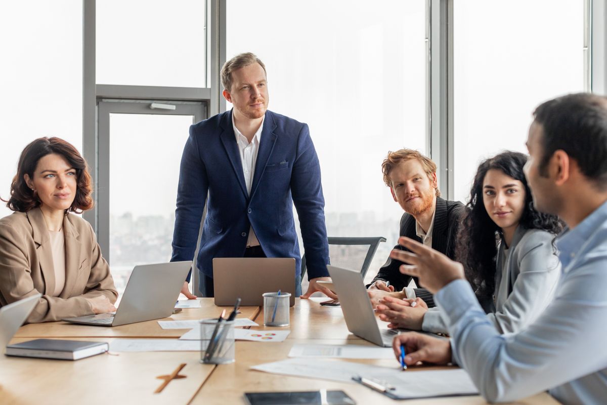 Egy jó álláshoz ma jobban kell a kreativitás és motiváltság, mint a sok diploma. A group of business professionals are gathered around a large wooden table in a modern office space. The men and women are engaged in a meeting, discussing ideas and strategies