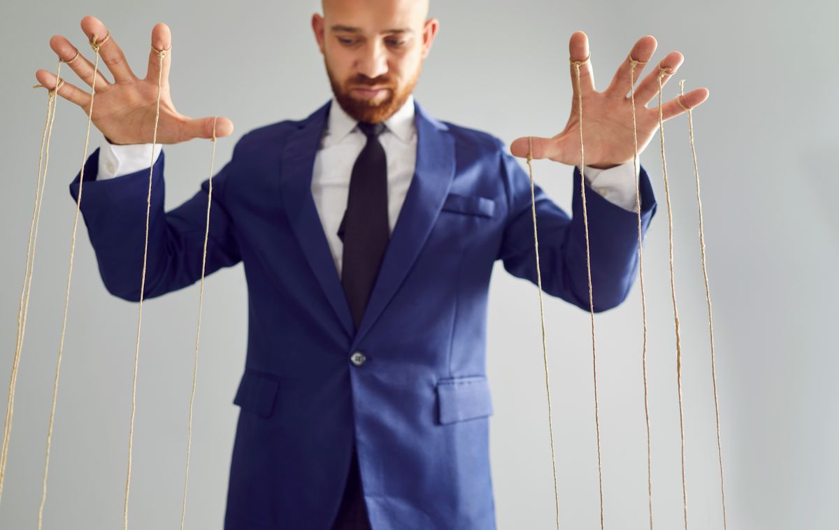 Öltönyös ember, aki bábmesterként szerepel a képen, kezéről madzagok lógnak.