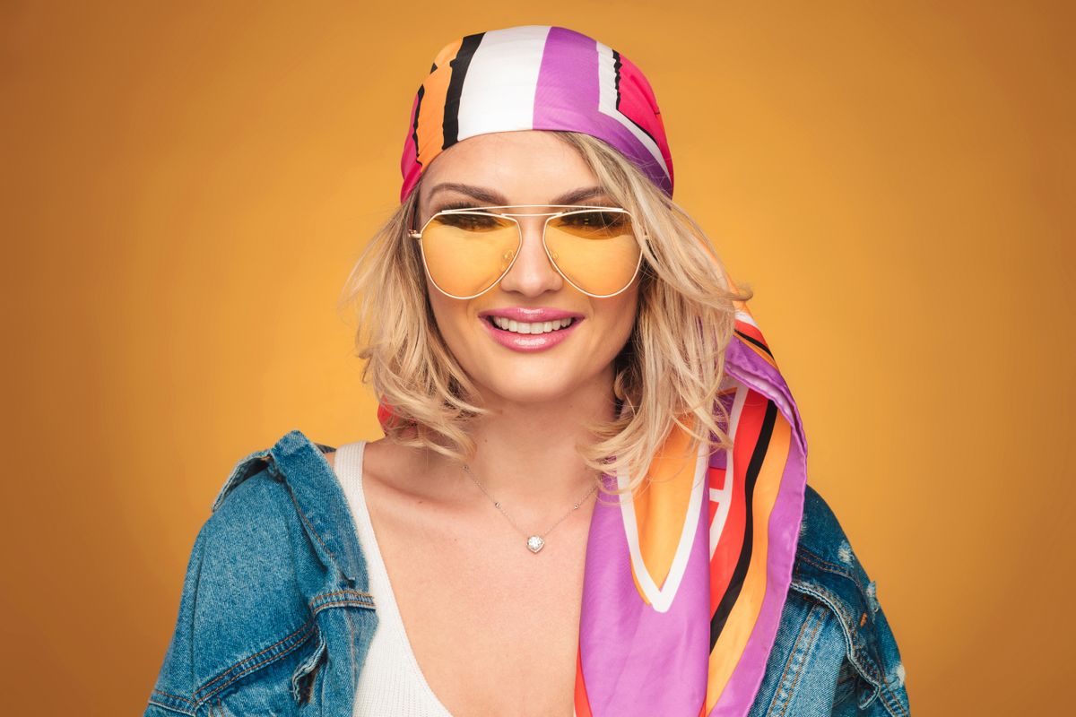 portrait of beautiful blonde woman with colorful outfit smiling on yellow background. She is wearing a colorful headscarf, a pair of yellow sunglasses and a blue undone denim shirt