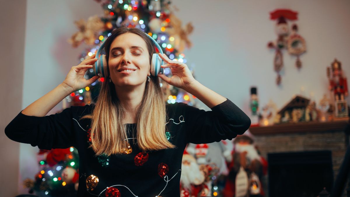Karácsonyi dalok: egy nő a karácsonyfa előt fülhallgatóval hallgatja a dalokat..