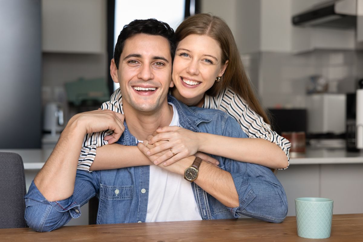 Cheerful attractive young Hispanic couple posing for portrait at home, hugging at kitchen table, looking at camera with toothy smiles, enjoying being together in comfortable apartment