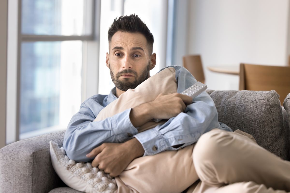 Sensitive Latin guy watching romantic love drama movie with emotional sympathy on home sofa, holding TV remote control, hugging pillow about to cry, feeling depressed, lonely