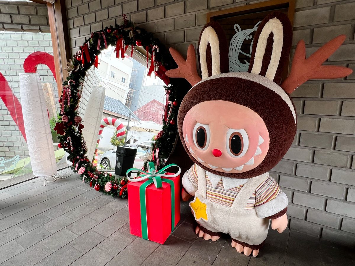 Rénszarvasnak öltözött karácsonyi labubu egy  nagy tükör mellett áll.