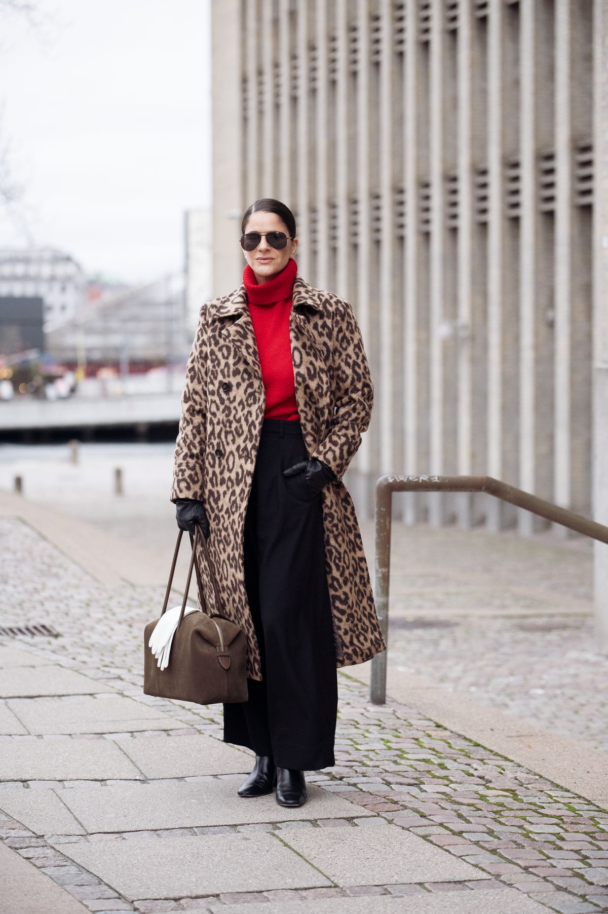 COPENHAGEN, DENMARK - DECEMBER 06: Gili Biegun wears leopard long coat by Baum und Pferdgarten, red knitted top, large Demellier London The Stockholm bag in Hazel suede with hanging white leather gloves, knitted black trousers and black leather gloves on December 06, 2025 in Copenhagen, Denmark. (Photo by Raimonda Kulikauskiene/Getty Images)