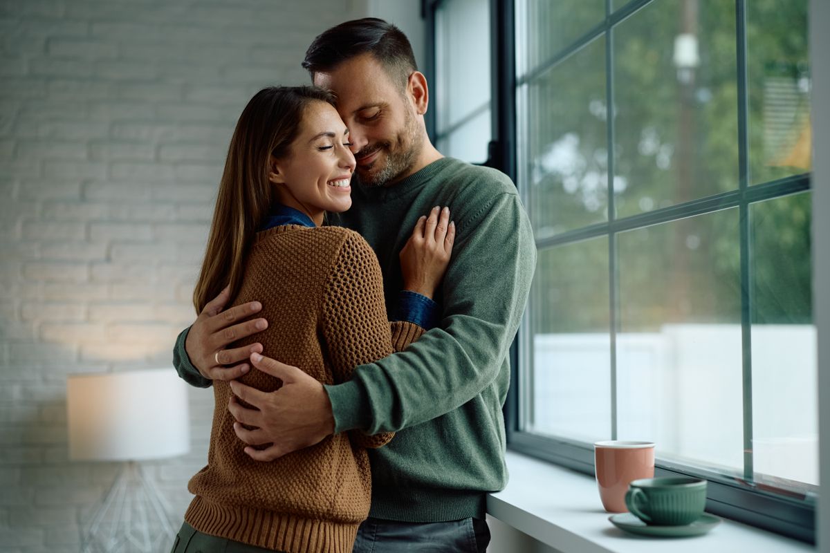 Smiling couple in love embracing by the window at home. Copy space.