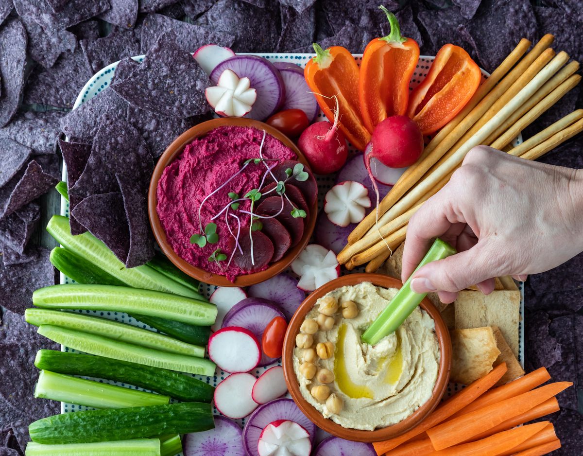 Close up of a hand dipping a celery stick into hummus dip with various colourful vegetable and blue corn chips surrounding.