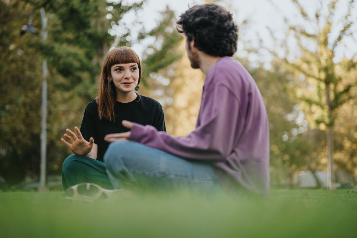 Two young people engage in a friendly conversation while sitting on the grass outdoors. The scene reflects friendship, connection, and relaxation in a green, sunlit environment.