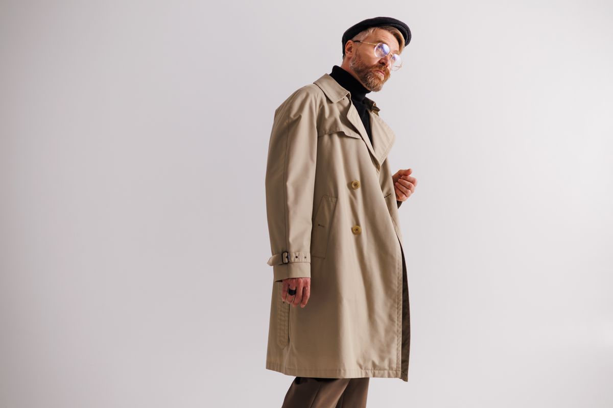 Fashionable blonde man in trench coat, eyeglasses and flat cap posing isolated on grey