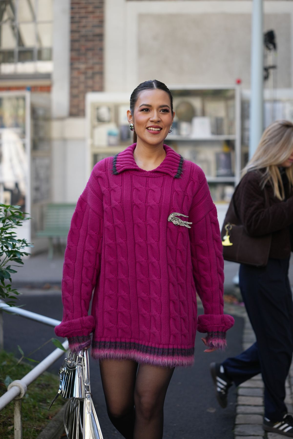 PARIS, FRANCE - OCTOBER 5: Raisa Adriana wears a silver and diamonds large earring, a pink purple wool oversized pullovers worn as a dress from Lacoste, a silver and rhinestones large crocodile brooch from Lacoste, silver large and small varnished leather pleated pattern handbags from Lacoste, black tights, outside Lacoste, during the Womenswear Spring Summer 2026 as part of Paris Fashion Week on October 5, 2025 in Paris, France. (Photo by Edward Berthelot/Getty Images)