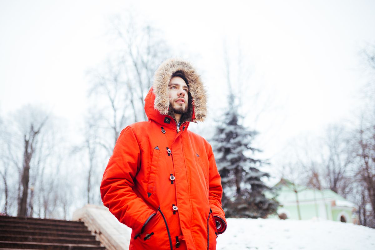 Portrait of a young stylish man with beard dressed in red winter jacket with hood and fur on his head stands against the backdrop of a snow-covered city and looks thoughtful. Winter and frost theme.
