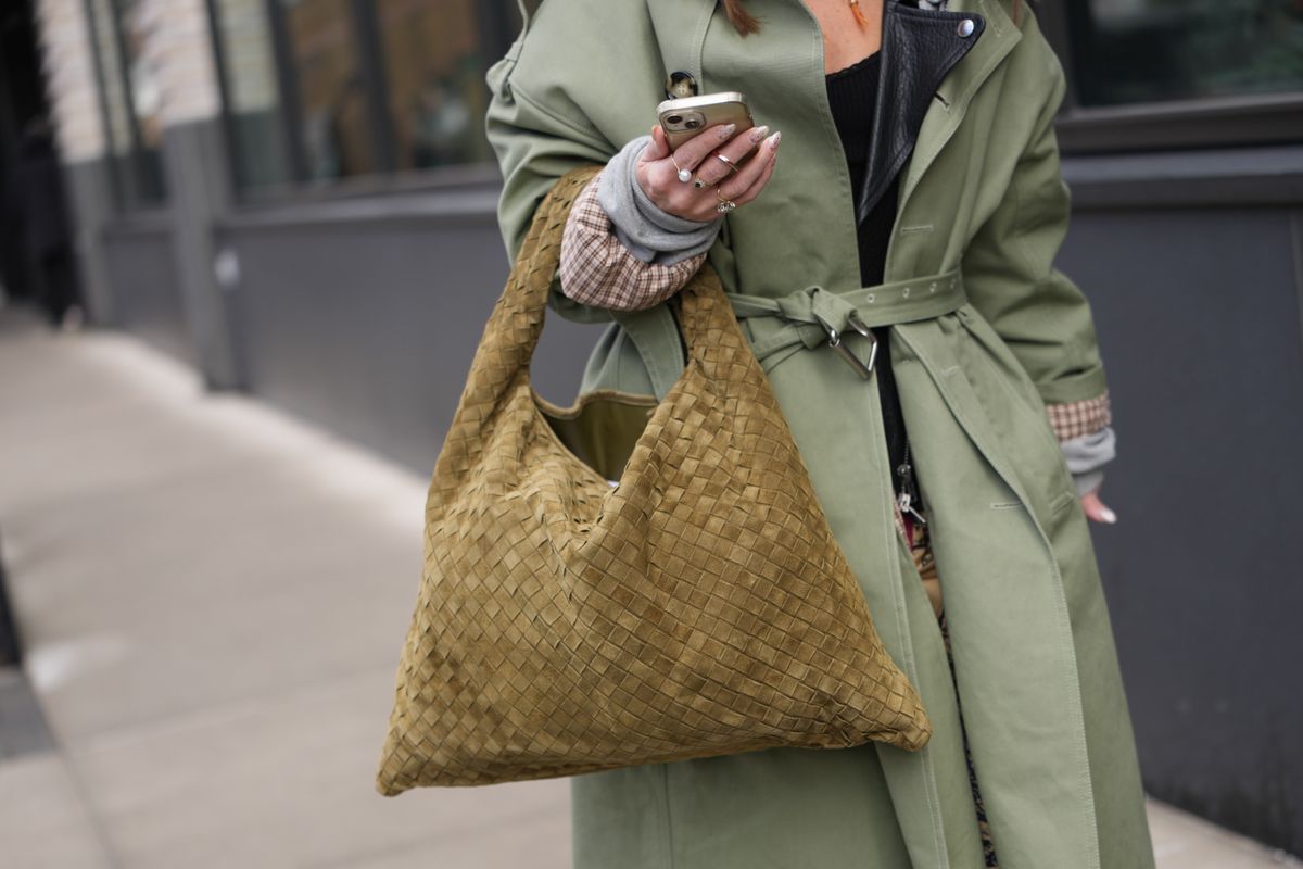NEW YORK, NEW YORK - FEBRUARY 12: A guest wears an olive woven suede oversized handbag from Bottega Veneta, a green belted trench coat, a black top, outside TWP, during New York Fashion Week, on February 12, 2026 in New York City (Photo by Edward Berthelot/Getty Images)