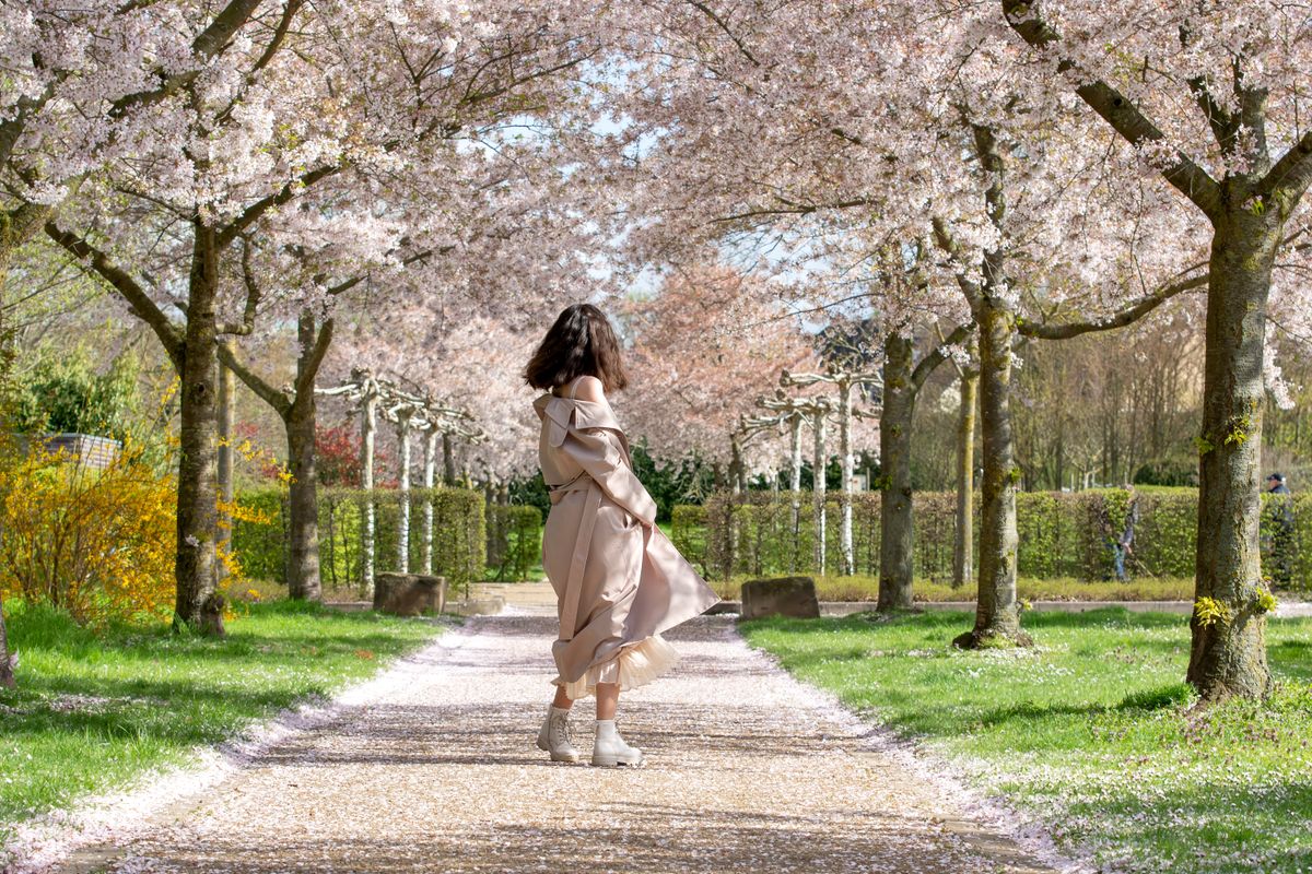 tavaszi kabát divat, illusztrációképp egy tavaszi fás környezetben táncol ballonkabátban egy fiatal nő, háttal a kamerának.
