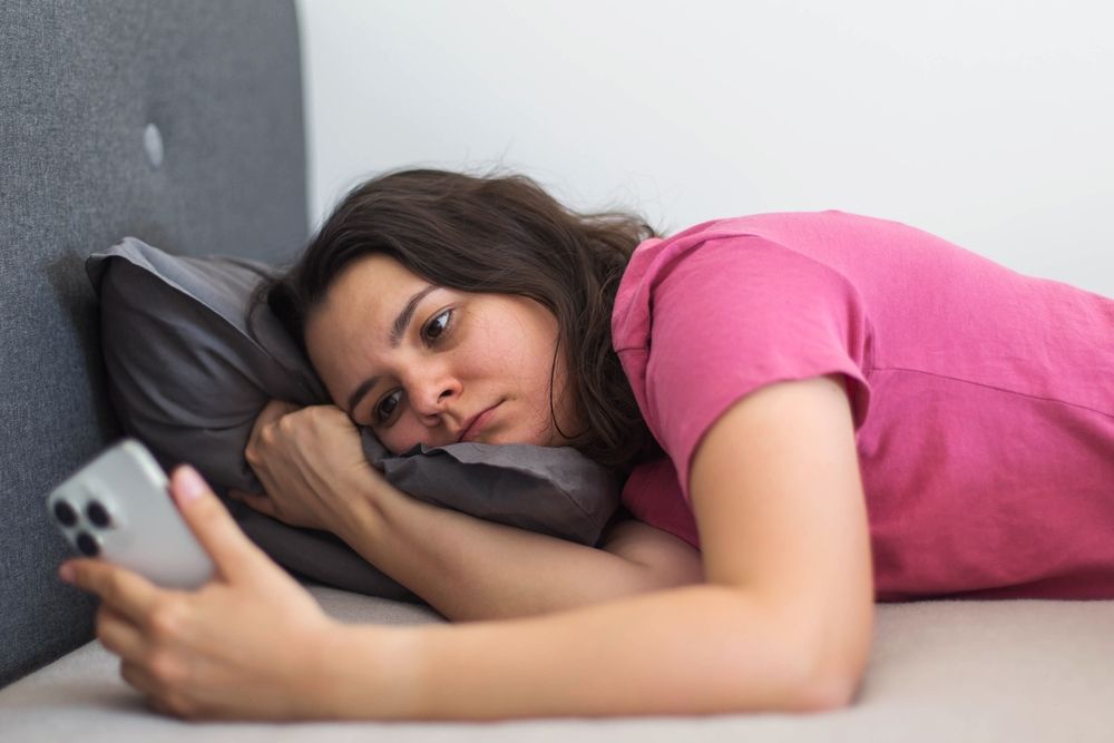 Unatkozó nő felszik az ágyon a telefonját bámulva. 