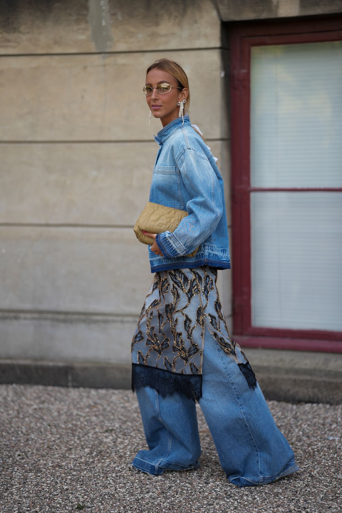 COPENHAGEN, DENMARK - AUGUST 6: Guiomar Machaz attending the Munthe show during Copenhagen Fashion Week SS26, is seen wearing a light-wash denim Munthe blouse, called Luxa Top, with a stand, mandarin-style, collar and half-placket closure, patch chest pockets, and a boxy, cropped silhouette; a pair of full-length, wide-leg light blue denim trouser by Munthe, called Lecube Pants, with a double hem at the end; overlaying the denim she is wearing a black transparent tulle Munthe dress, called Lazo dress, with an all-over gold floral pattern with metallic gold thread and a wide lace trim at the bottom; she carries a Roluke bag by Munthe, a soft leather bag in khaki with a quilted floral pattern; her blond hair is slicked back; she wears a pair of slim, oval metal-frame glasses; a pair of chandelier earrings composed of cascading faux pearl beads that form a ribbon shape during Copenhagen Fashion Week day three on August 6, 2025 in Copenhagen, Denmark.  (Photo by Moritz Scholz/Getty Images)
