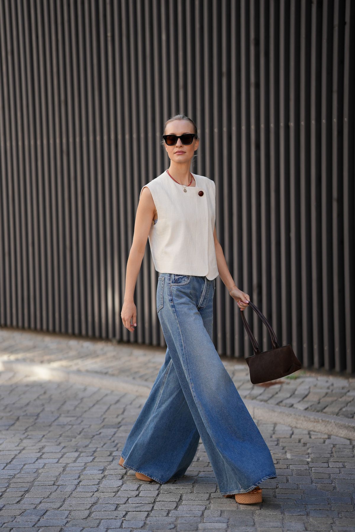 MUNICH, GERMANY - JULY 18: Marlies Pia Pfeifhofer is seen wearing a cream white sleeveless textured linen wrap top with asymmetric front panel and a large brown button closing at the shoulder from Sézane; light blue washed ultra wide leg Paloma jeans in structured denim with high waist and long flared silhouette from Citizens of Humanity; beige Dolly Verona raffia platform sandals with thick sole from Larroudé; a dark brown suede The New York shoulderbag with top handles and triangular fold design from DeMellier; square-shaped black sunglasses with thick acetate frame from CHIMI Eyewear; a surigao pearl necklace with red Japanese glass beads and a freshwater baroque pearl from Les Beads Studio; a golden medallion necklace with a faceted diamond bezel and a hand-painted 'Porte Bonheur' enamel inscription from Marlo Laz; golden mini hoop earrings from Monica Vinader; her blonde hair is styled in a sleek high bun on July 18, 2025 in Munich, Germany. (Photo by Moritz Scholz/Getty Images)