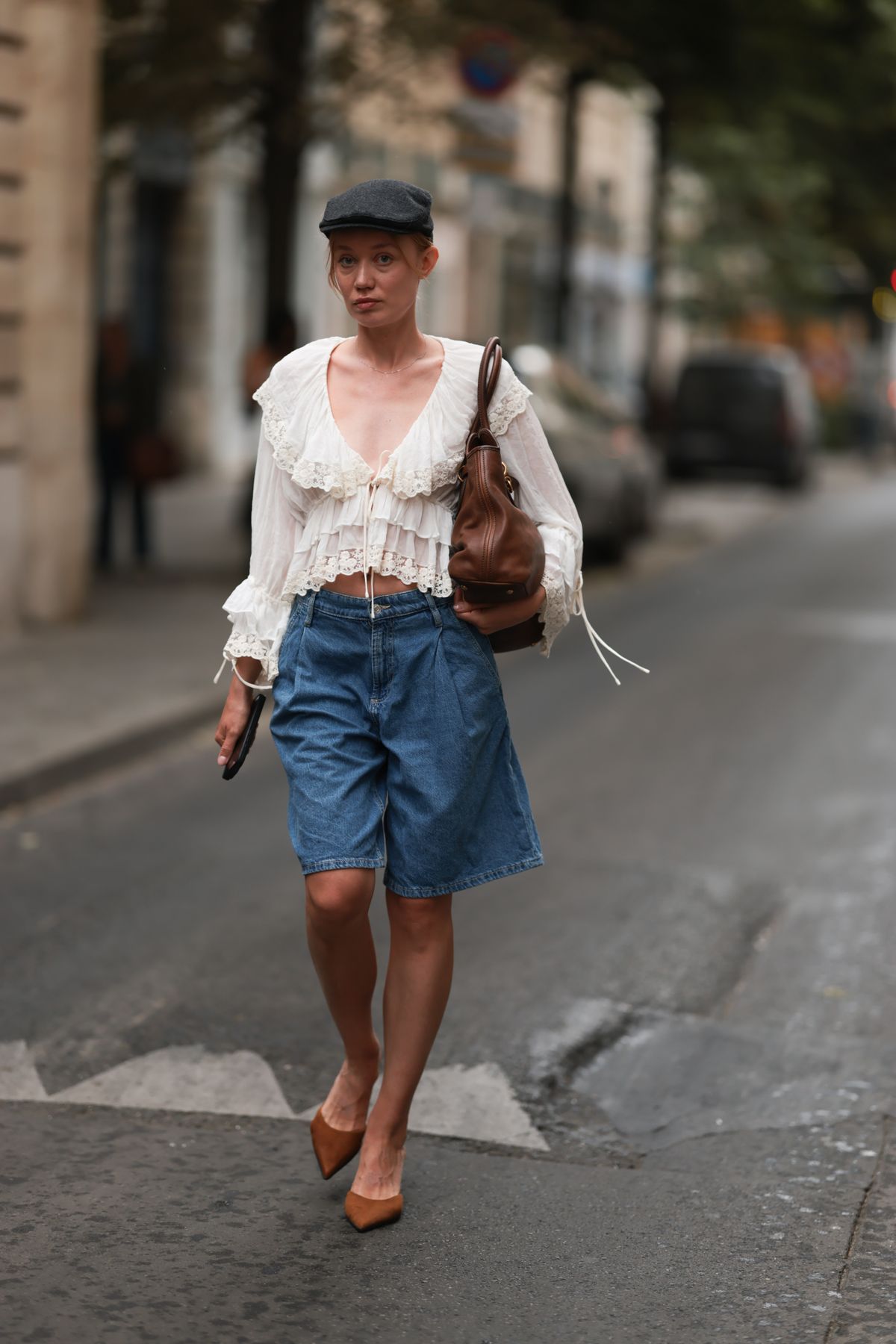 PARIS, FRANCE - JUNE 25: A Fashion Week guest is seen wearing a dark grey newsboy cap, a fine silver necklace, a cream-white long-sleeved ruffled blouse with a deep V-neck and lace details, a dark brown leather shoulder bag, wide-leg blue Bermuda denim shorts, and pointed brown suede mules with kitten heels, while holding an Apple iPhone during the Jeanne Friot Menswear Spring/Summer 2026 show as part of Paris Fashion Week on June 25, 2025 in Paris, France. (Photo by Jeremy Moeller/Getty Images)