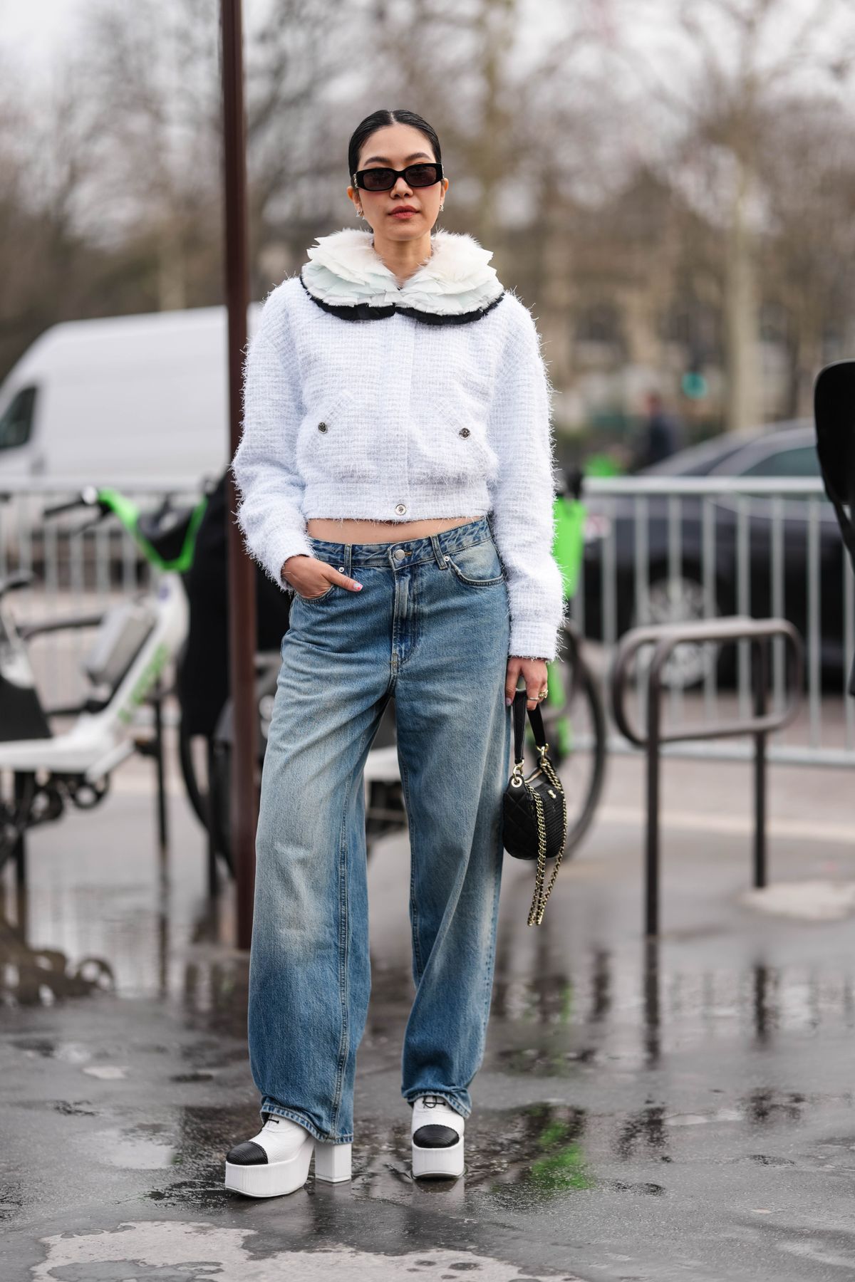 PARIS, FRANCE - MARCH 11: A guest wears black sunglasses, silver earrings, white tweed textured white light blue black feathered collar oversized Chanel bomber jacket, navy blue washed denim jean pants, shiny black Chanel leather bag, white platform heeled black toe heels Chanel leather shoes, outside Chanel, during the Paris Fashion week Women's Fall/Winter 2025-2026 on March 11, 2025 in Paris, France. (Photo by Edward Berthelot/Getty Images)