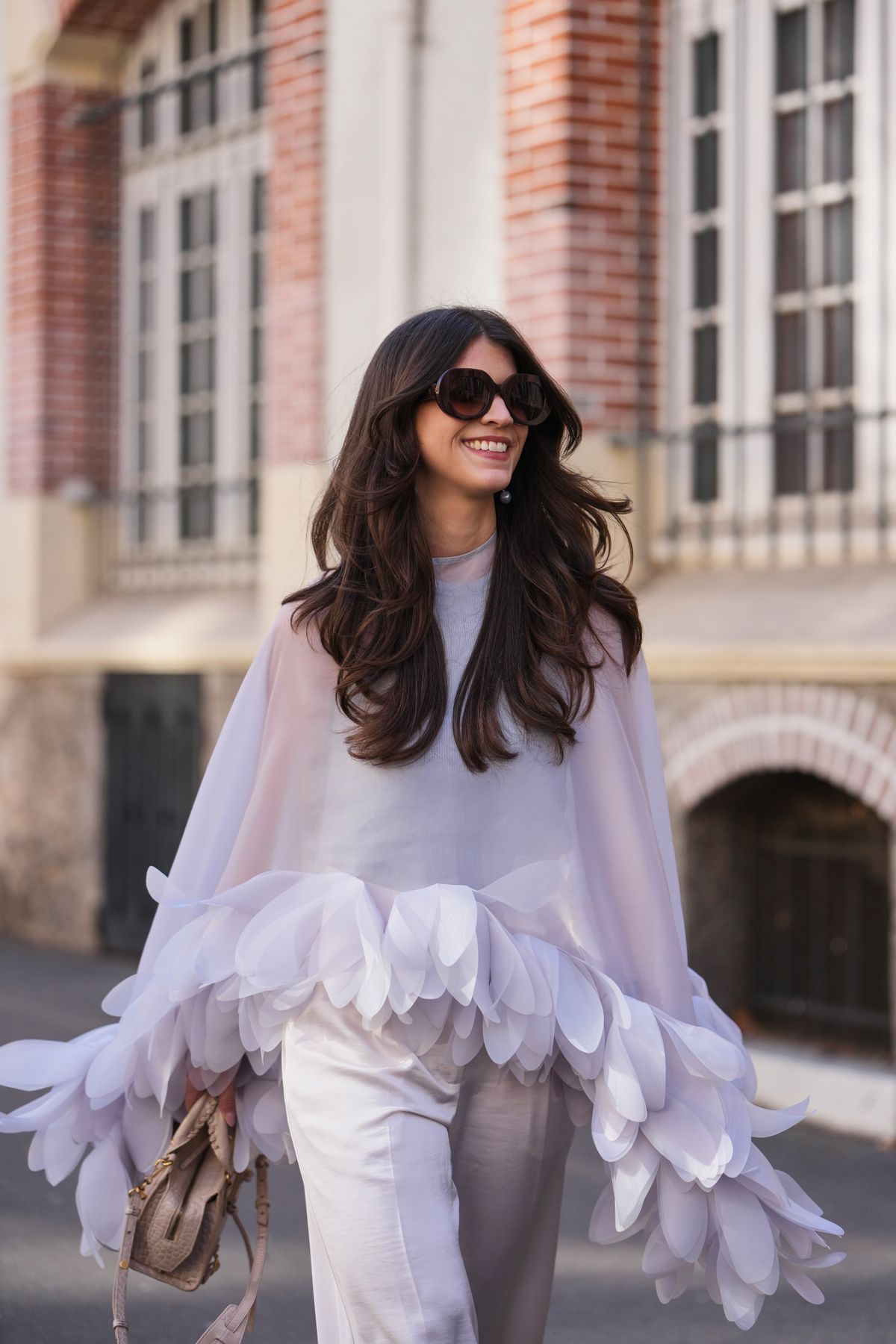 PARIS, FRANCE - MARCH 04: A guest wears brown wavy hair, black sunglasses, a light lavender ruffled sheer cape with white layered petal trim, white satin wide leg pants, a beige leather bag, outside Dries Van Noten, during Paris Fashion Week - Womenswear Fall/Winter 2026/2027, on March 04, 2026 in Paris, France (Photo by Edward Berthelot/Getty Images)