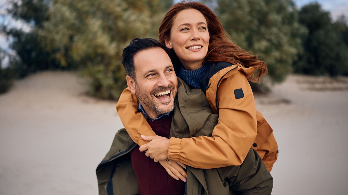 nőiesség megélése, párkapcsolati teszt: egy szerelmes pár kedvesen mosolyog a kamerába, a nő a férfi hátán van