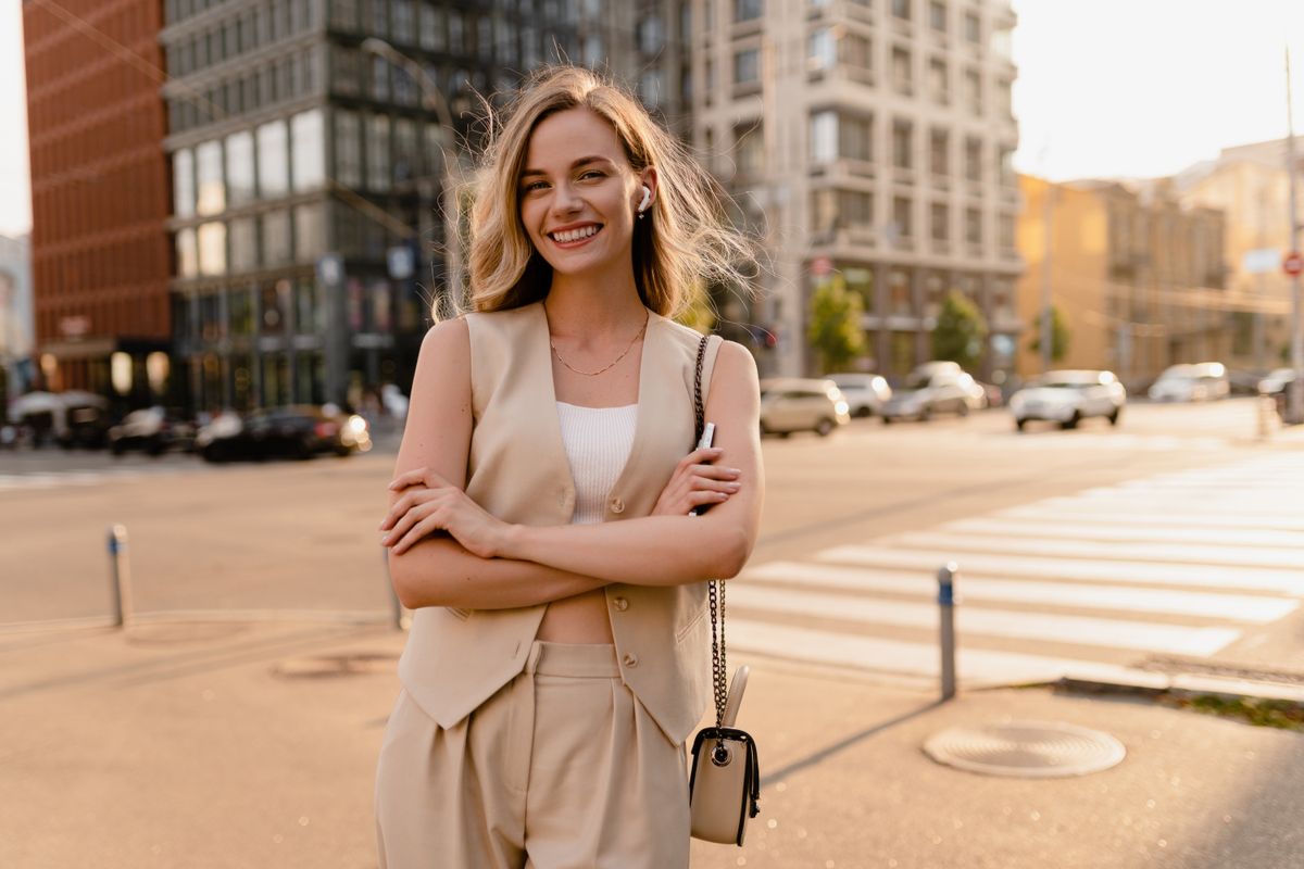 pretty blonde woman walking in sunny city street in beige suit with a purse in urban fashion style, smiling, summer trend season, wearing vest and trousers, glam hair hairstyle