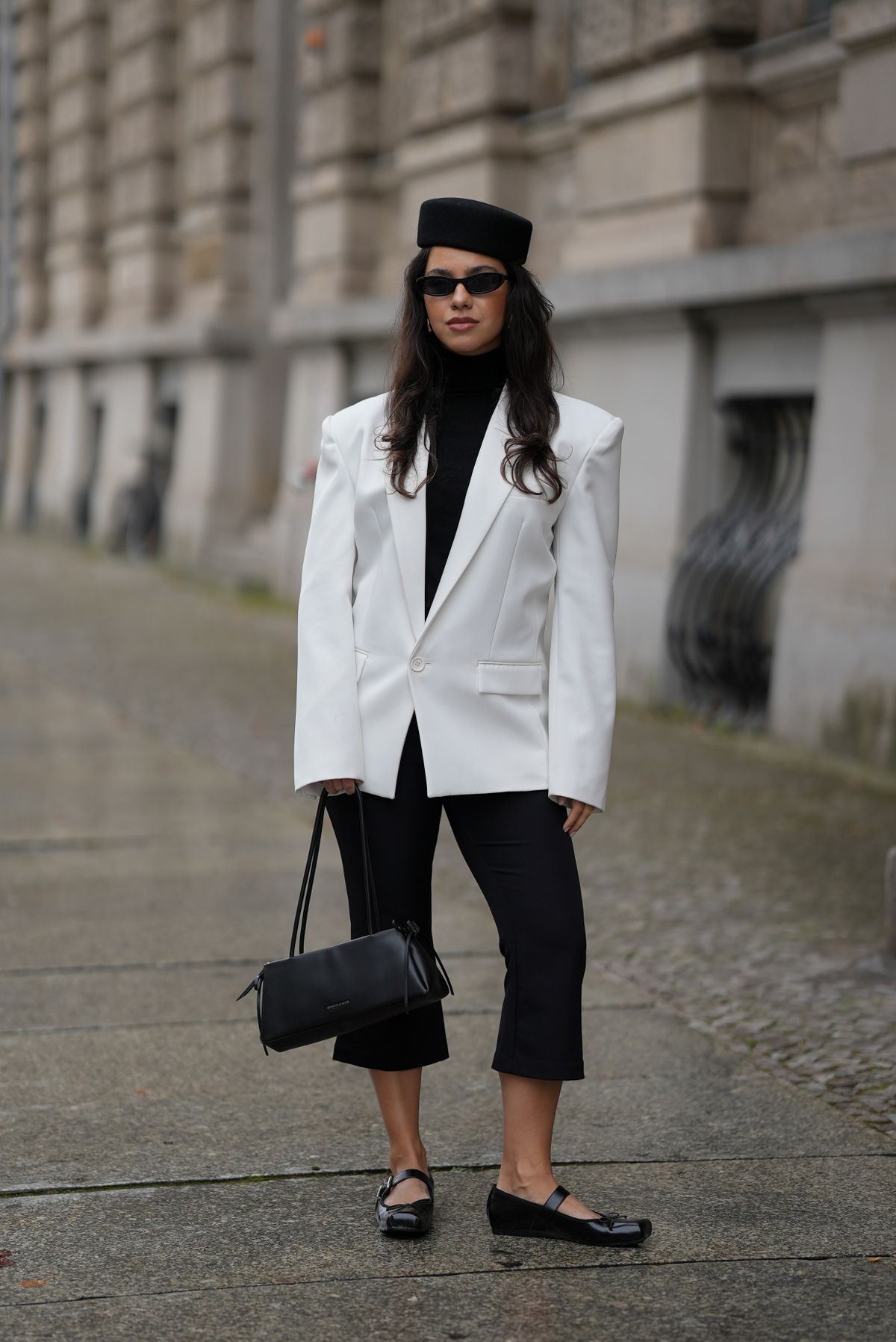 BERLIN, GERMANY - OCTOBER 22: Bella Emar is seen wearing a white oversized blazer with structured shoulders by The Frankie Shop; a fitted black turtleneck sweater by Massimo Dutti; cropped black capri pants with a slight flared cut by H&M; black patent leather Mary Jane ballerinas with square toes, buckle straps and bow details by Charles & Keith; a structured black leather purse with bow details by Charles & Keith; slim black sunglasses with a shiny frame by H&M; and a black wool pillbox hat called “Oriana” by Fae Nuit; her long dark brown hair is worn loose in soft waves on October 22, 2025 in Berlin, Germany. (Photo by Moritz Scholz/Getty Images)