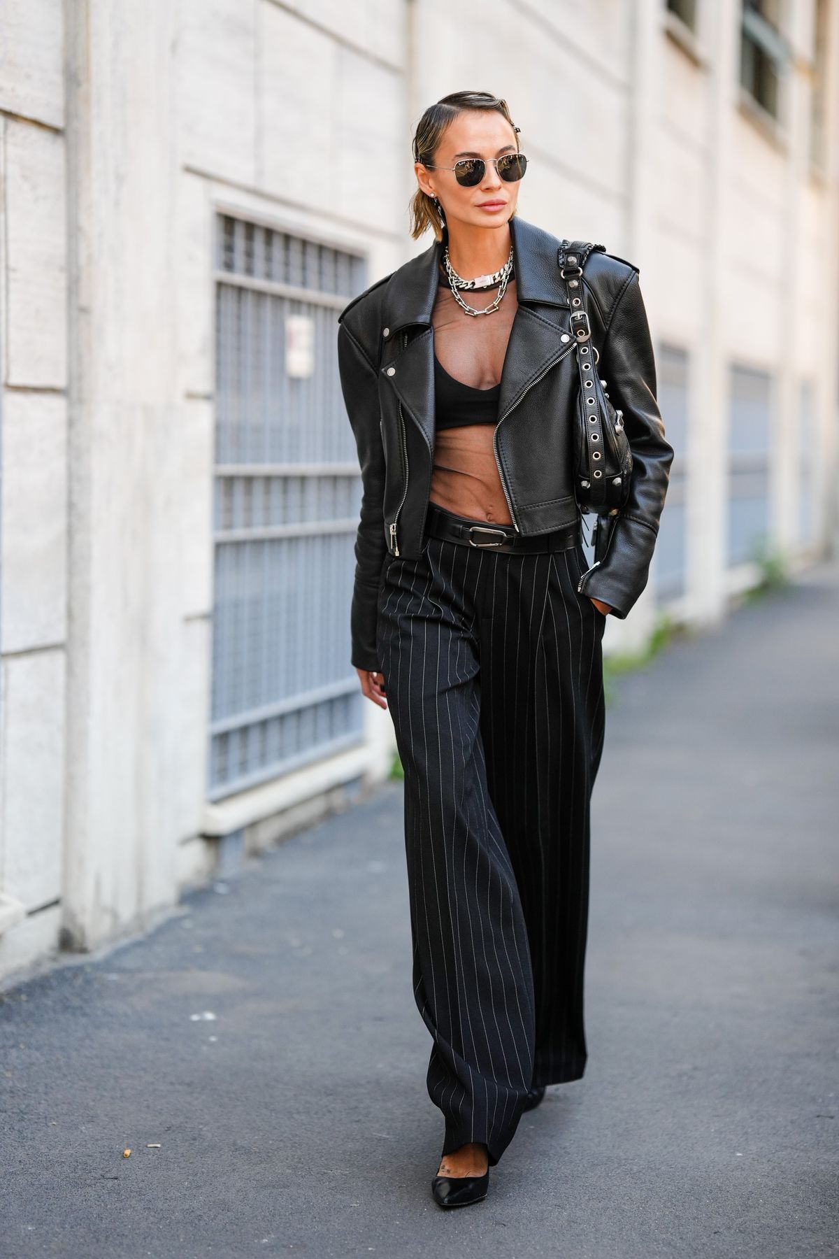 MILAN, ITALY - SEPTEMBER 22: A guest wears black sunglasses from Ray Ban, silver earrings, silver hair clips, a black transparent t-shirt, silver large chain necklace, a black bra underwear, a black shiny leather biker jacket, a black shiny leather belt, a black shiny leather nailed / studded shoulder bag from Balenciaga, black with gray stripes large palazzo pants, black shiny leather pointed pumps heels shoes, outside the Alberta Ferretti fashion show during the Milan Fashion Week - Spring / Summer 2022 on September 22, 2021 in Milan, Italy. (Photo by Edward Berthelot/Getty Images)