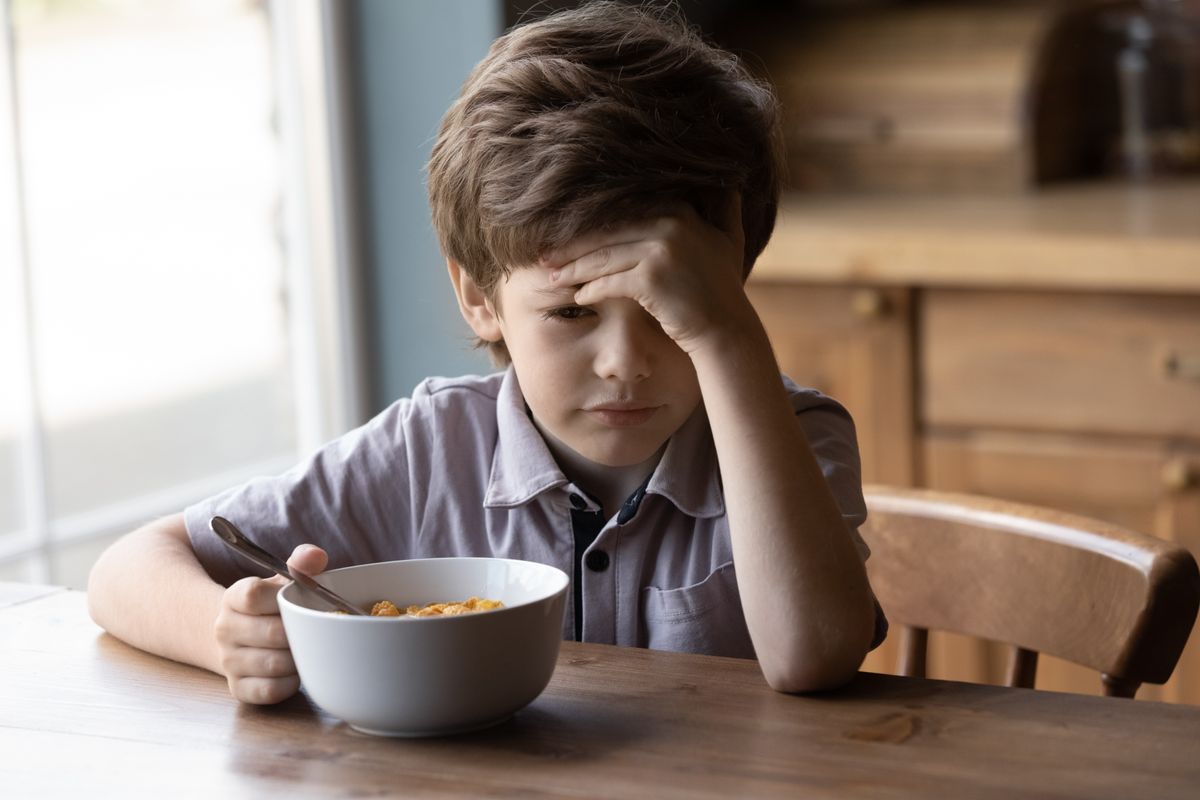 Unhappy little Caucasian boy child sit at table at home kitchen have no appetite for tasty healthy breakfast. Upset stressed small kid refuse to eat delicious organic cereals with milk. Diet concept.