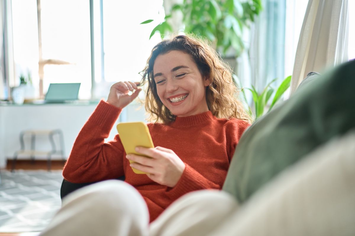 Happy relaxed young woman sitting on couch using cell phone, smiling lady laughing holding smartphone, looking at cellphone enjoying doing online ecommerce shopping in mobile apps or watching videos.