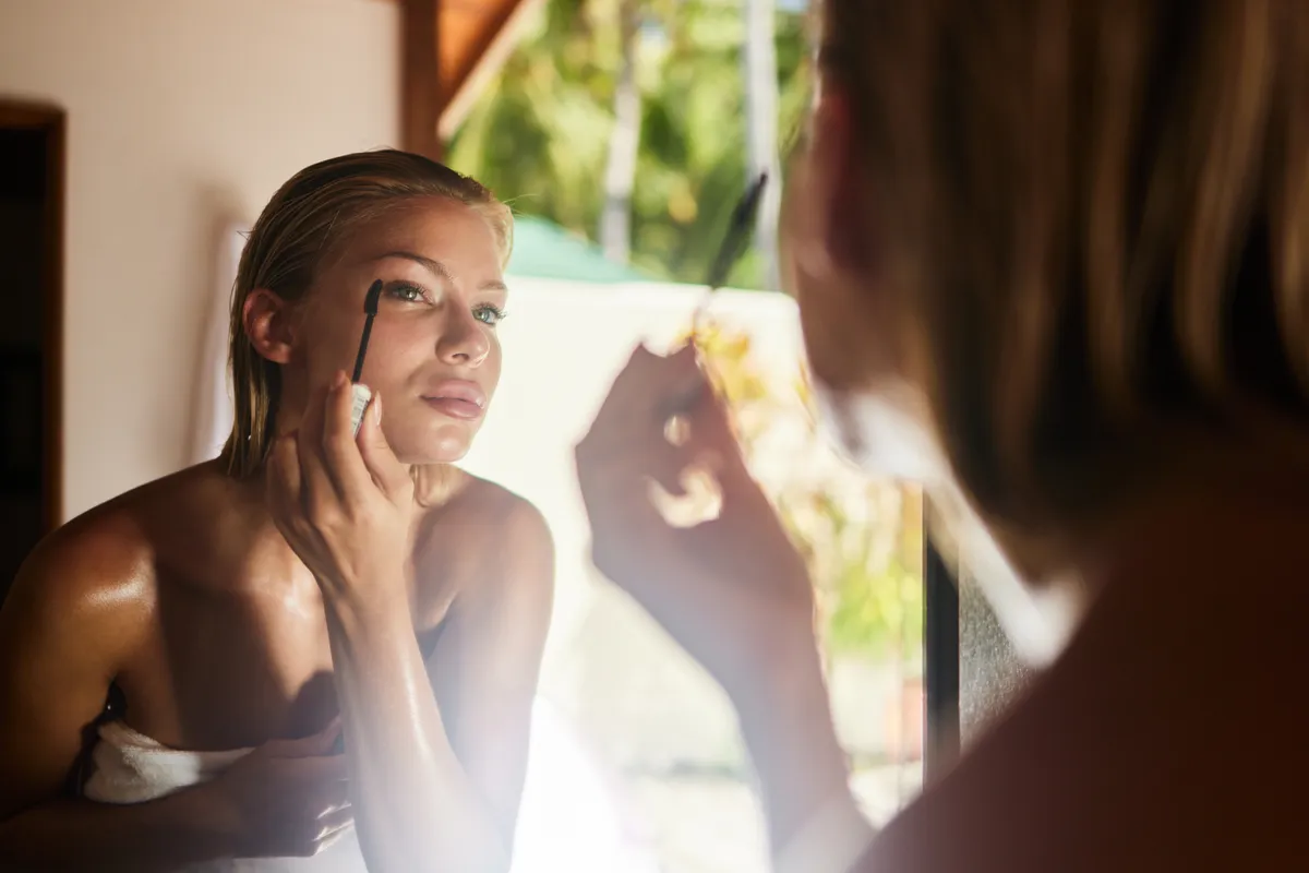 Reflection in a mirror of young beautiful woman applying mascara after the shower in bathroom.