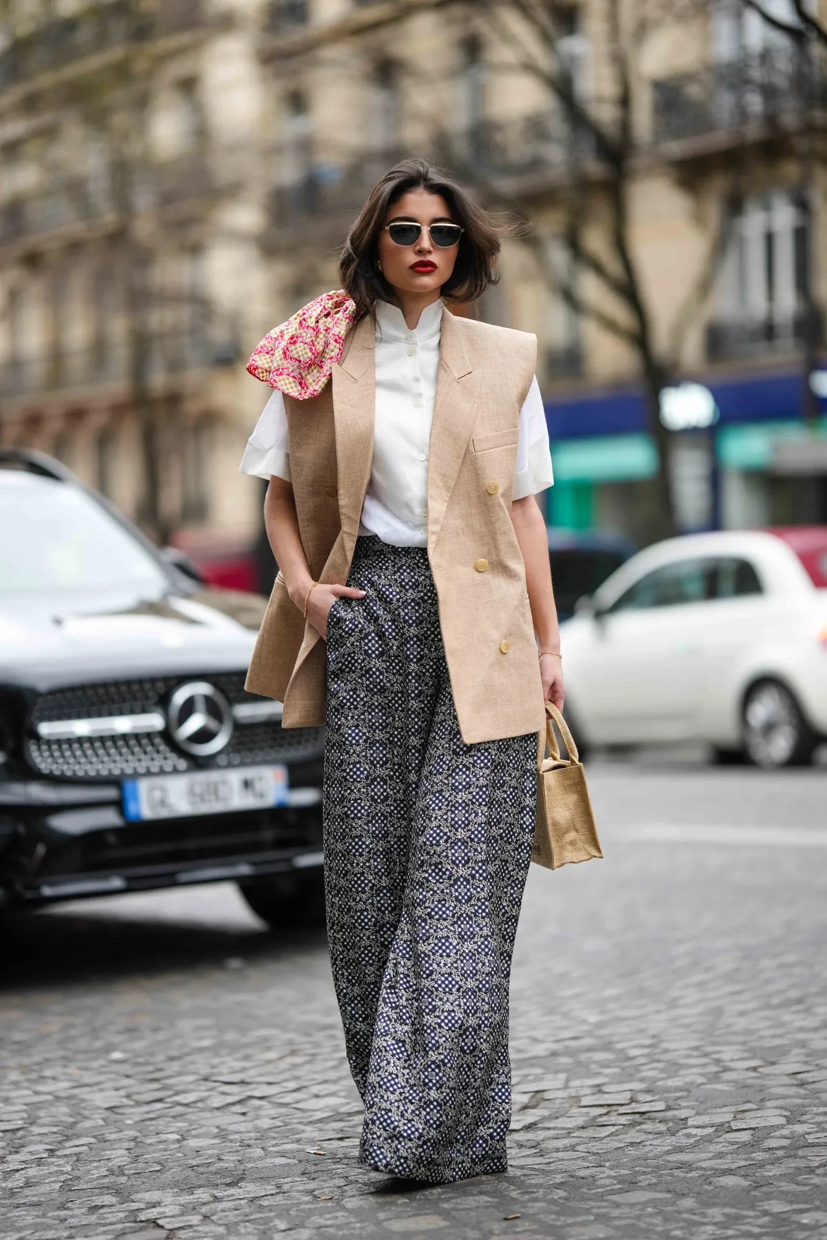 PARIS, FRANCE - MARCH 05: A guest wears black and gold sunglasses, a white high neck buttoned blouse short sleeves shirt, a beige linen sleeveless blazer gilet, a beige wicker handbag, a white and black print pattern large flowing pants, pumps heels shoes , outside EENK, during Paris Fashion Week - Womenswear Fall Winter 2023 2024, on March 05, 2023 in Paris, France. (Photo by Edward Berthelot/Getty Images)